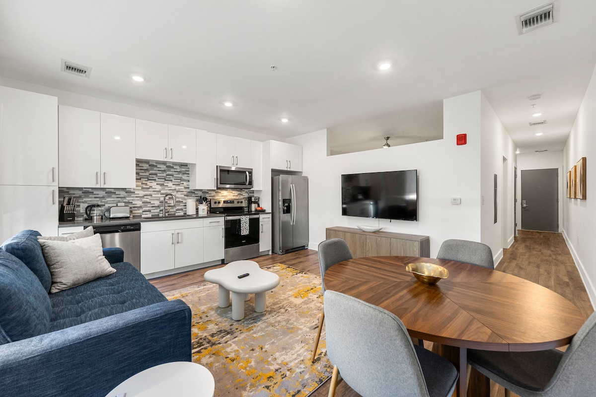 The open-plan living area features a modern kitchen with sleek cabinetry, stainless steel appliances, and a stylish backsplash. A comfortable blue sofa is positioned adjacent to a rounded coffee table, while a circular dining table for four is set near the flat-screen TV.