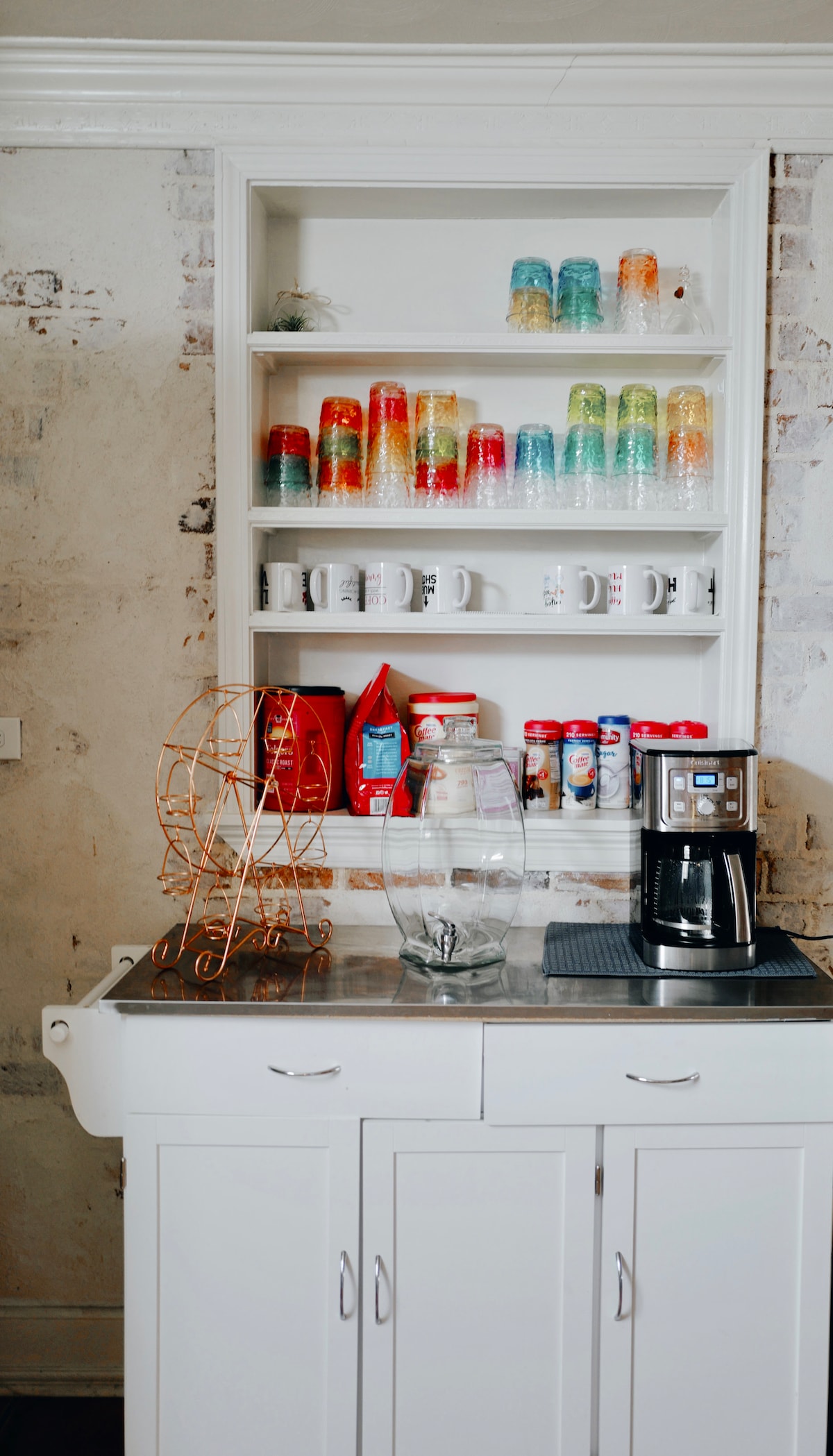 A well-organized kitchen shelf displays an array of colorful glassware and mugs alongside various coffee condiments. A coffee maker rests on a stainless steel surface, with a decorative glass jar nearby, contributing to a functional kitchen space.
