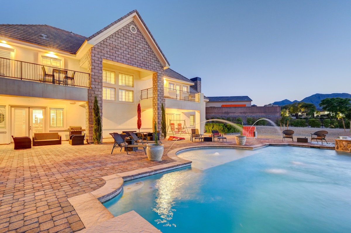 The exterior of the estate features a spacious patio with lounge seating and an outdoor dining area beside a shimmering pool. Water cascades gently from a fountain into the pool, with mountain views visible in the background under a twilight sky.