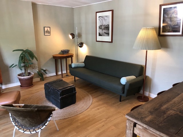 A retro-style living area features a green sofa with rounded armrests, accompanied by a wooden side table and a floor lamp. A circular woven rug adds warmth to the space, complemented by a dark chest serving as a coffee table. Potted greenery enhances the inviting setting.