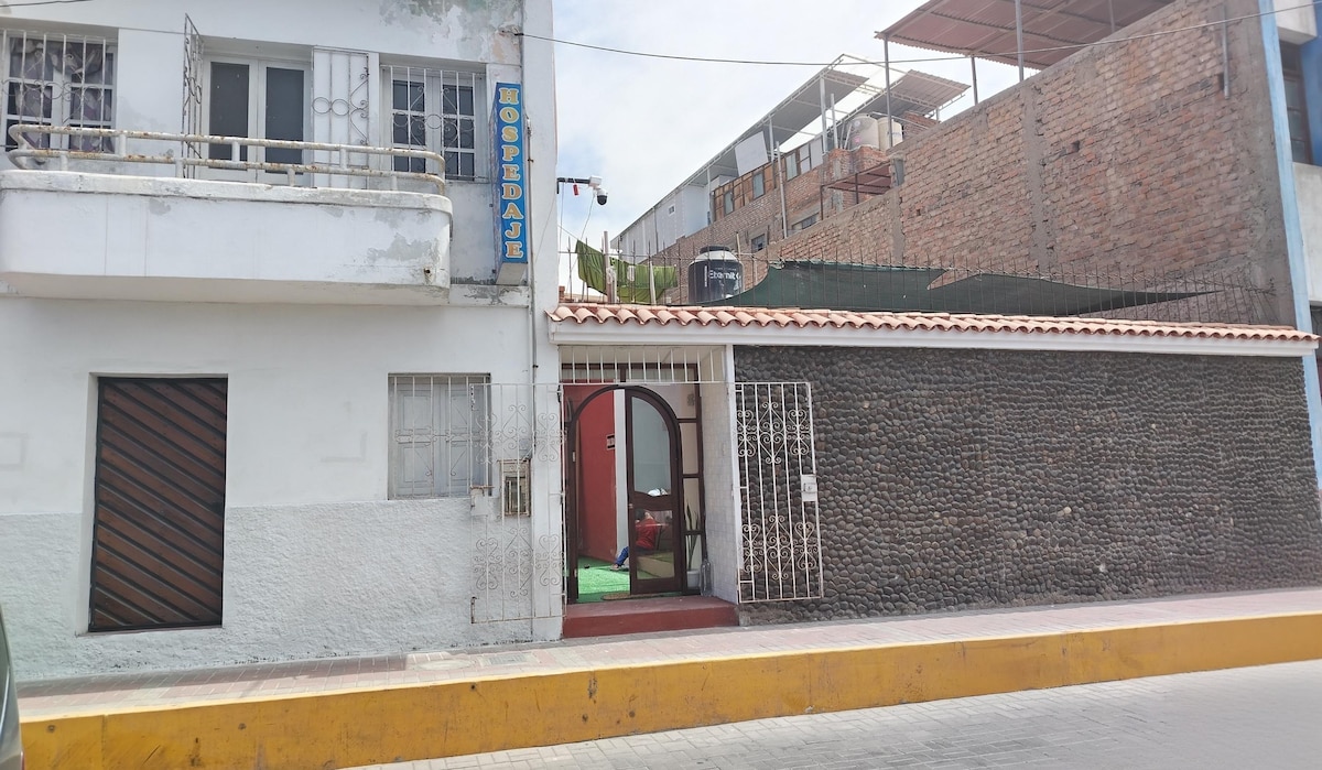 The exterior of a guesthouse is depicted, featuring a white building adjacent to a stone wall. An arched entrance is visible, leading to a green space. The sign above indicates the accommodation, while surrounding structures display varying materials and textures.