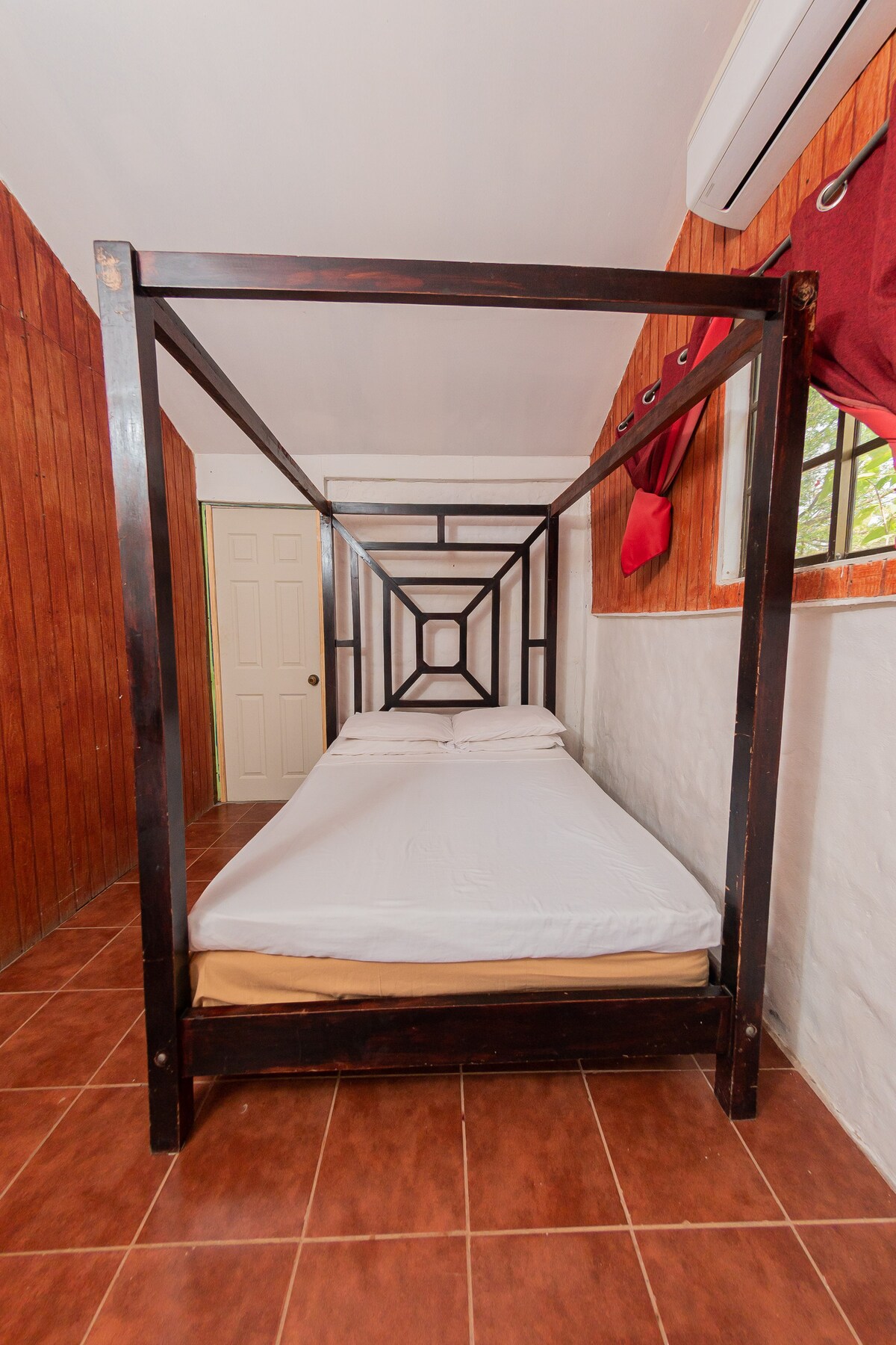 A cozy sleeping space showcases a canopy bed framed in dark wood, featuring a simple white bedding set. The walls are adorned with warm wooden panels, accompanied by red curtains that frame the window, allowing natural light to enter the room.