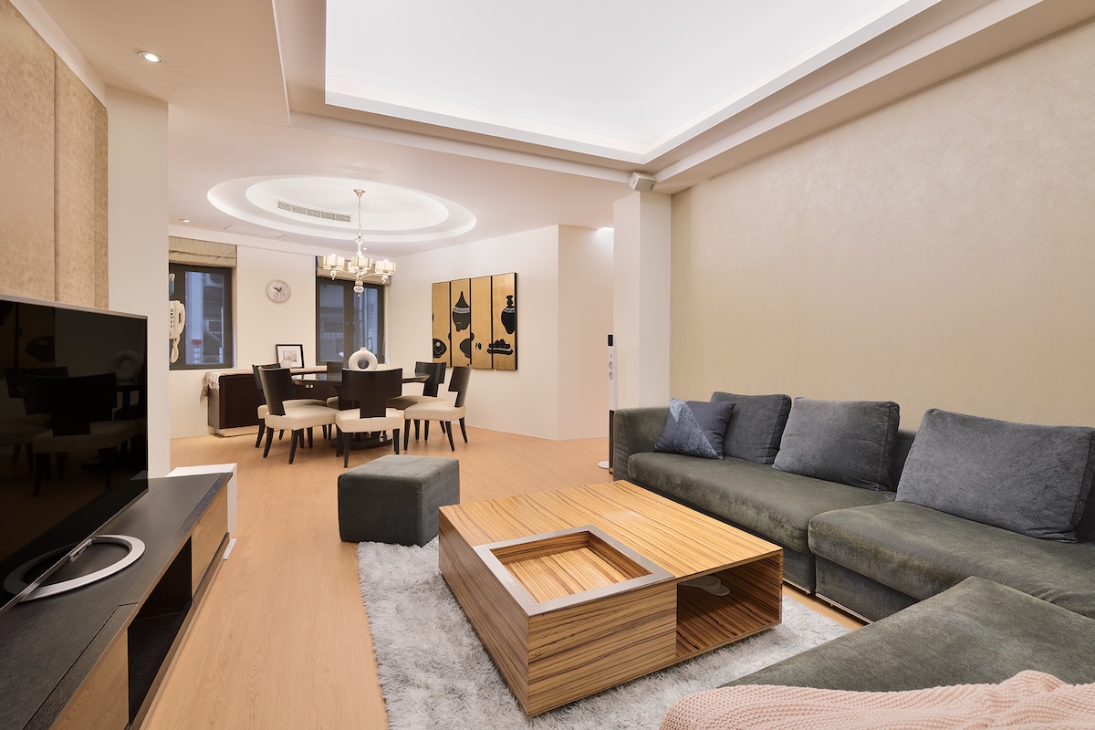 The living area features a sectional sofa and modern coffee table placed on a soft area rug. A dining table is seen in the background, accompanied by dining chairs. Soft, ambient lighting creates a welcoming atmosphere, while large windows provide natural light.