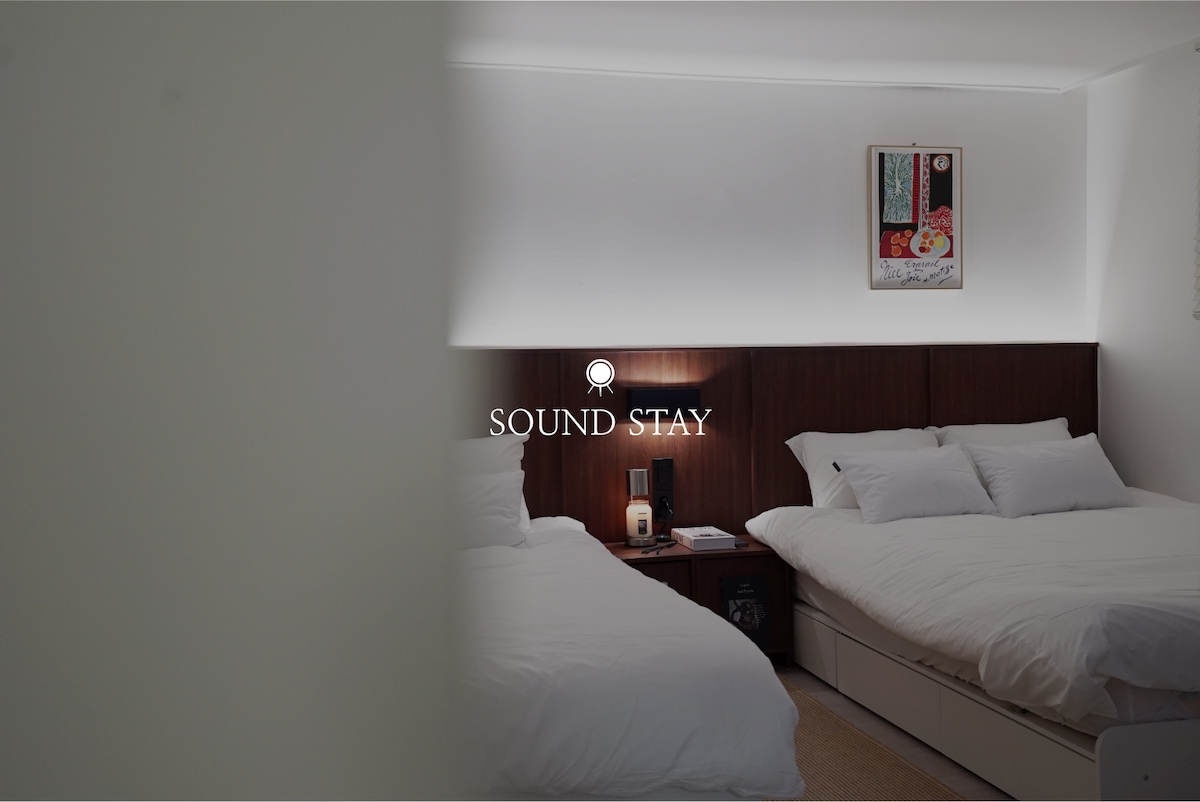 A spacious bedroom featuring two hotel-grade queen beds dressed in crisp white linens. A modern bedside table with a lamp is positioned between the beds. Soft lighting highlights the minimalist decor, and a piece of framed art decorates the wall.