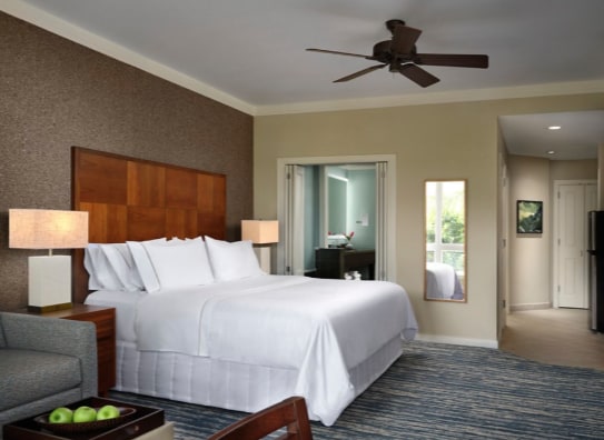 A spacious studio villa features a king bed with white bedding, complemented by bedside tables with lamps. A comfortable seating area is present, along with a full-length mirror. Light-colored walls and carpet add to the inviting ambiance, while a ceiling fan is installed for air circulation.