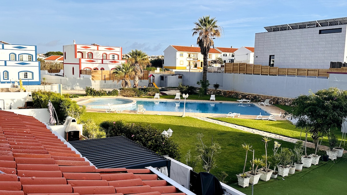 A spacious outdoor area is showcased, featuring a large swimming pool surrounded by greenery and lounge chairs. Palm trees line the poolside, and buildings with colorful exteriors are visible in the background. Clear skies enhance the overall bright and inviting setting.