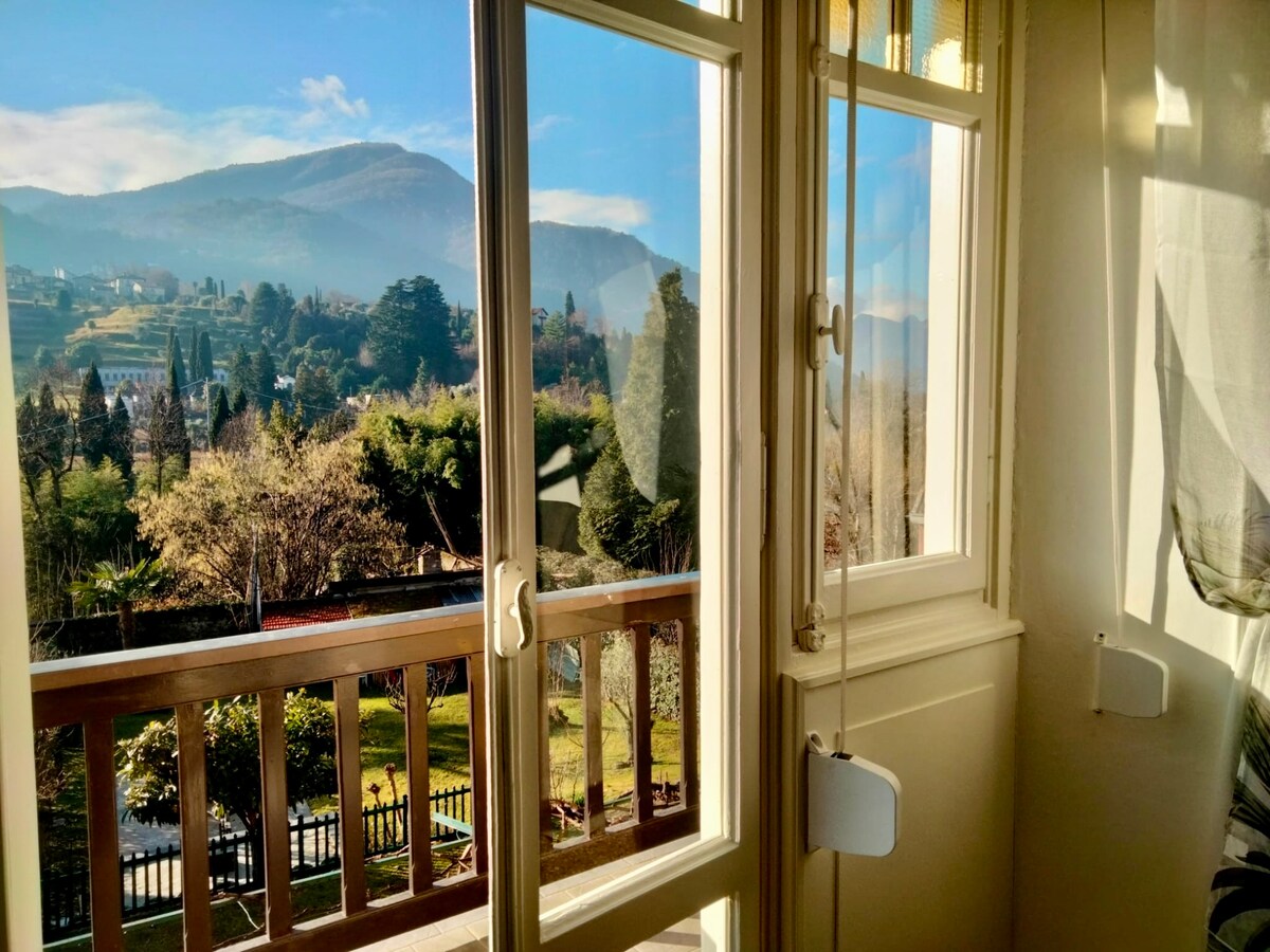 A view from a window frames the lush greenery of the garden and distant mountains. Bright natural light filters through the glass, highlighting the serene outdoor landscape. The railing adds a touch of elegance to the indoor space, seamlessly connecting it with the scenery.