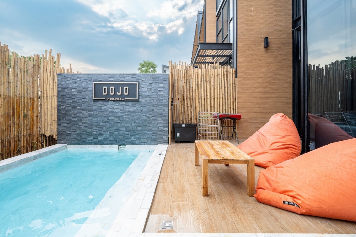 A private pool area is surrounded by bamboo privacy fencing, featuring a light blue pool and a wooden table. Two vibrant orange bean bags are placed nearby, inviting relaxation. The structure behind showcases a modern design with large glass panels allowing natural light.