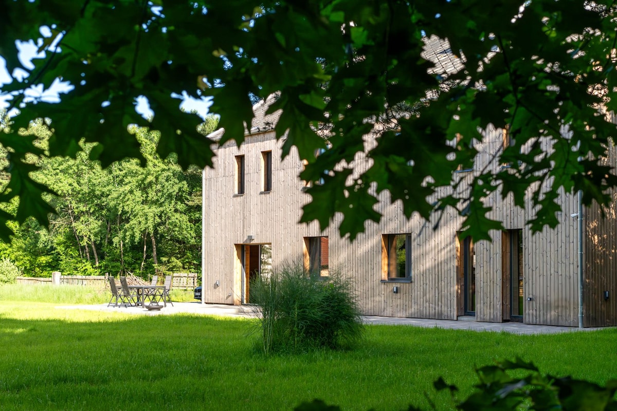 The image displays a wooden structure surrounded by lush greenery, illuminated by natural sunlight. A neatly maintained lawn enhances the serene environment, while garden furniture is visible, inviting guests to relax outdoors. The setting features a tranquil atmosphere, complemented by trees framing the view.