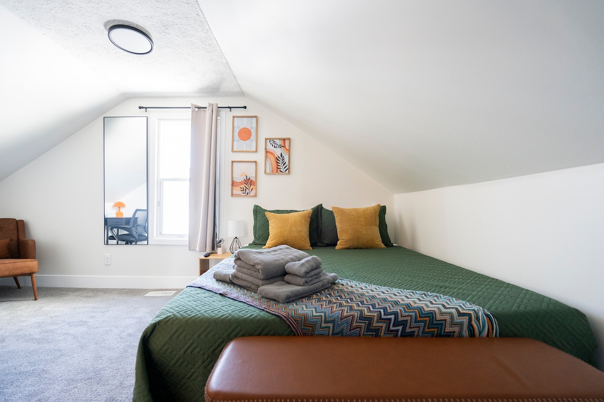 A cozy private bedroom features a spacious green bed, accented with yellow pillows. A decorative throw and neatly folded towels are placed at the foot of the bed. A comfortable chair is positioned nearby, and bright artwork adorns the walls, enhancing the room's inviting atmosphere.