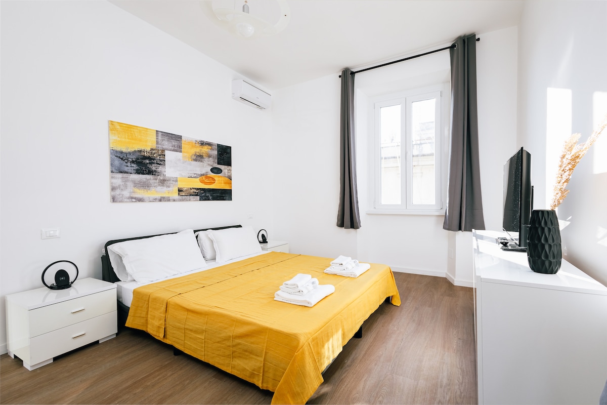 A spacious bedroom features a large bed with a yellow coverlet and two white pillows. Flanking the bed are two nightstands with black lamps. A dresser is positioned against the wall, and a television rests atop it. Natural light enters through the window, which is framed by grey curtains.