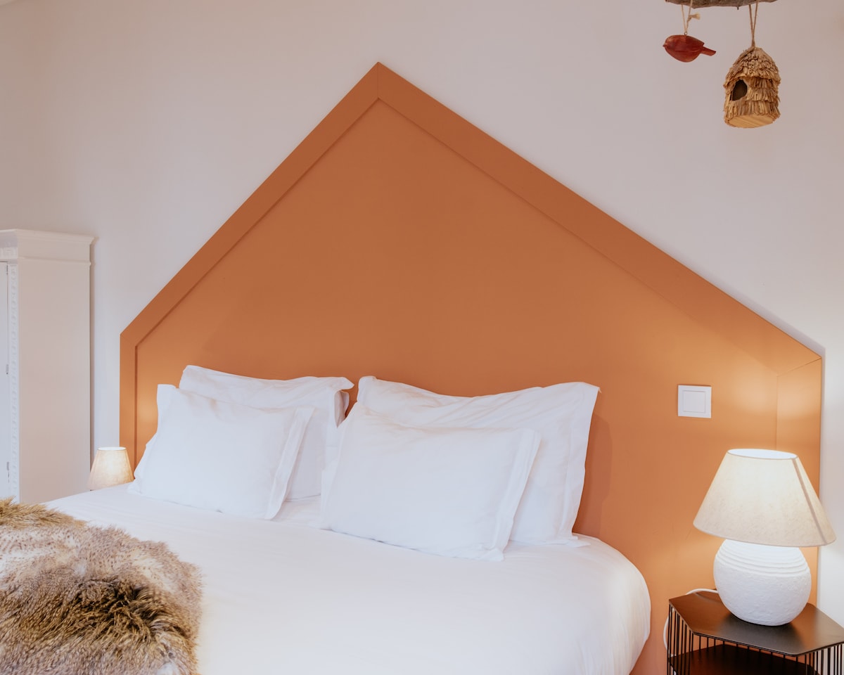 A spacious bed is centered against an orange accent wall, characterized by a distinctive gabled shape. Two white pillows and a soft blanket rest on the bed. A softly glowing lamp on a nearby nightstand adds gentle illumination to the cozy sleeping area.