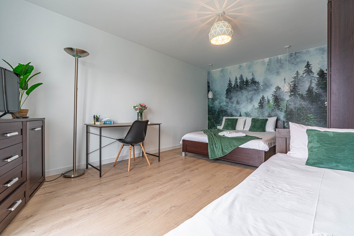 A modern bedroom features two spacious beds covered with crisp white linens and green accents. A wooden desk with a chair is situated beside a mountain-themed wall mural. A tall plant and a dresser enhance the room's functionality, while a floor lamp provides additional lighting.