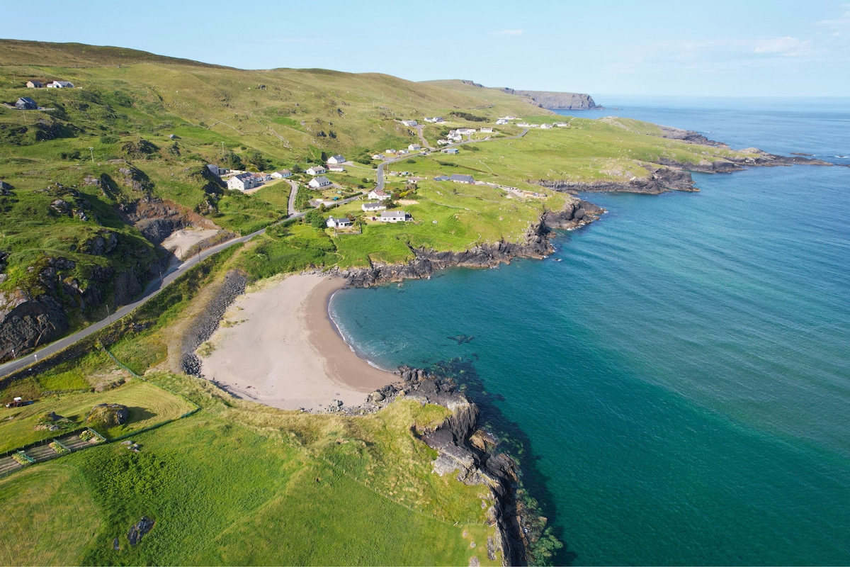 An aerial view captures a serene beach bordered by lush green hills. The gentle waves of the ocean lap at the sandy shore, while a few quaint homes are visible along the coastline. The expansive landscape offers a sense of tranquility and spaciousness.