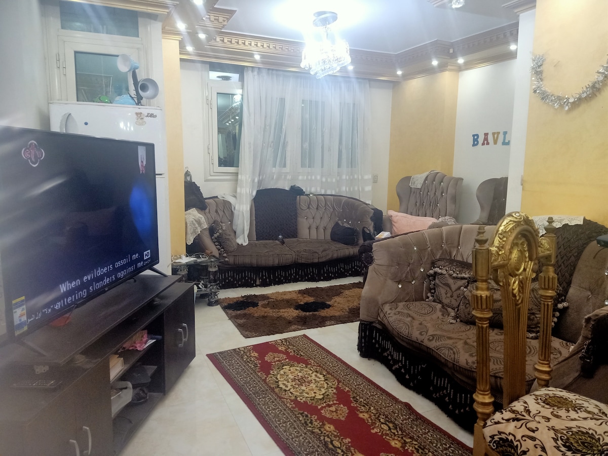 The living area features an arrangement of plush seating, highlighted by ornate patterns and warm colors. A large television is positioned on a dark wood cabinet, while a decorative area rug adds texture to the tiled floor. Soft lighting is provided by a chandelier overhead.