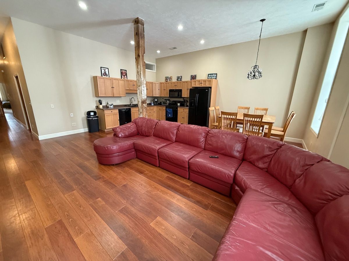 A spacious living area features a large red sectional sofa, which accommodates multiple guests. The open layout includes a dining table set for six and an adjoining kitchen, equipped with modern appliances. Natural light fills the room through generous windows.