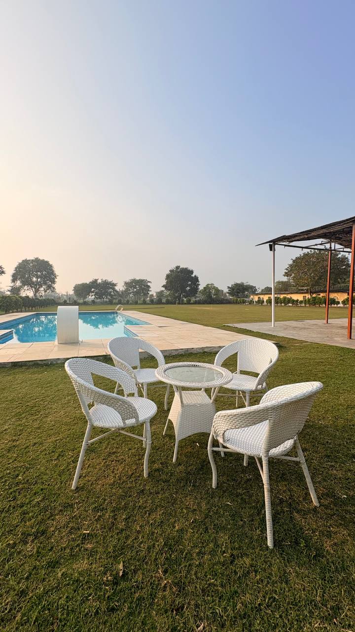 A serene outdoor seating area is set by a sparkling pool. Five white wicker chairs surround a glass-top table, positioned on lush green grass. The tranquil landscape features trees in the background, contributing to a peaceful ambiance.