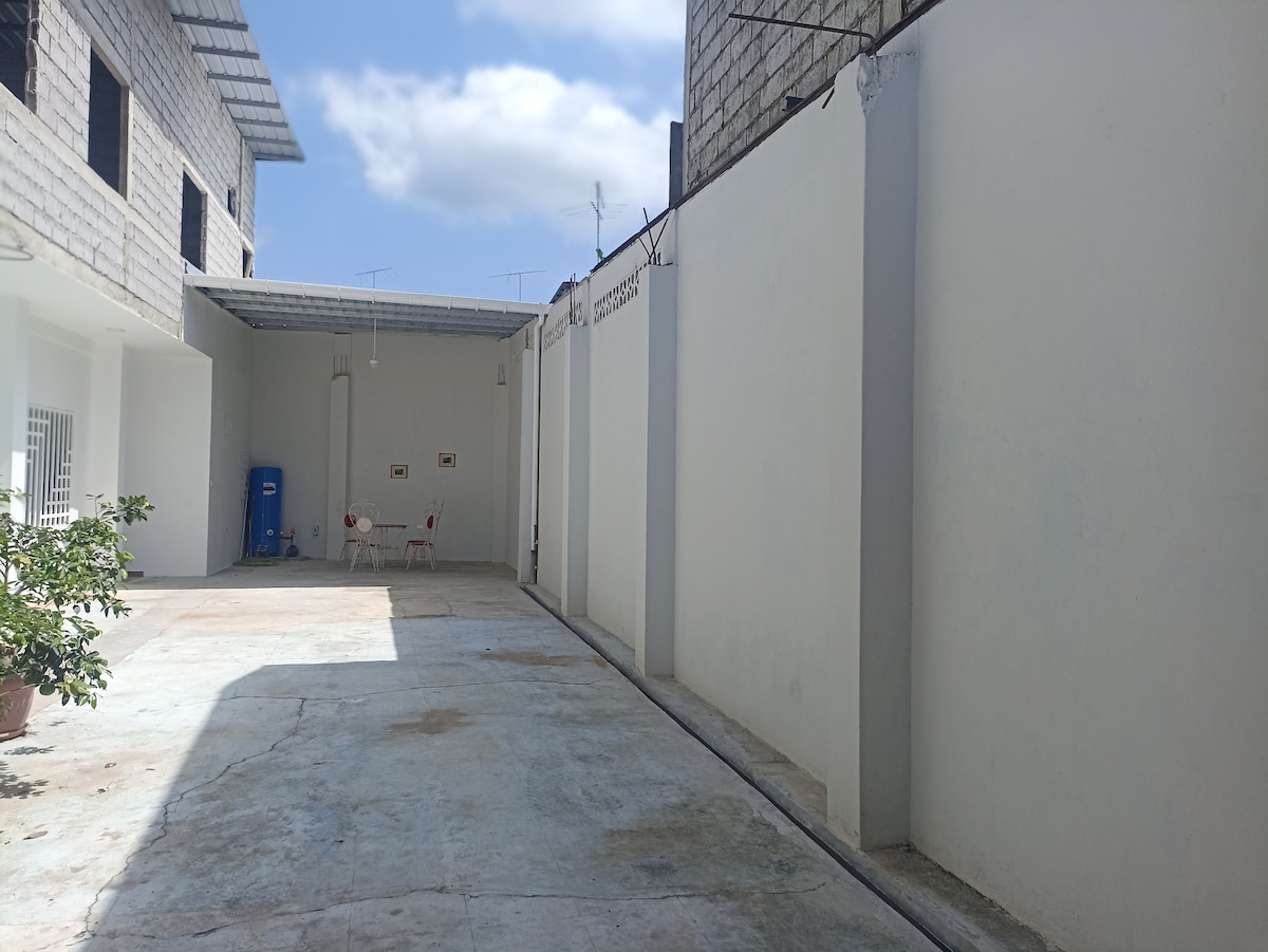 A spacious outdoor area is visible, featuring a concrete floor and white walls. Two red chairs are positioned against one wall, providing a place to relax. A blue water dispenser is situated in the corner, and sunlight filters in, creating a bright atmosphere.