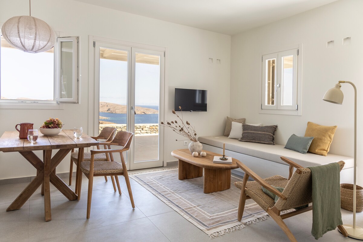 A minimalist living area features a wooden dining table with four chairs. A comfortable seating area includes a sofa with neutral-colored cushions and a low coffee table. Expansive windows offer views of the Aegean Sea, while a light fixture hangs above the space.