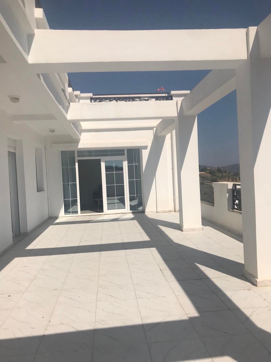 A spacious terrace area features clean, white marble flooring. Large glass doors lead to the interior, while shadows from the architectural elements create interesting lines across the surface. The surrounding railings offer a sense of openness with views of the mountains in the background.