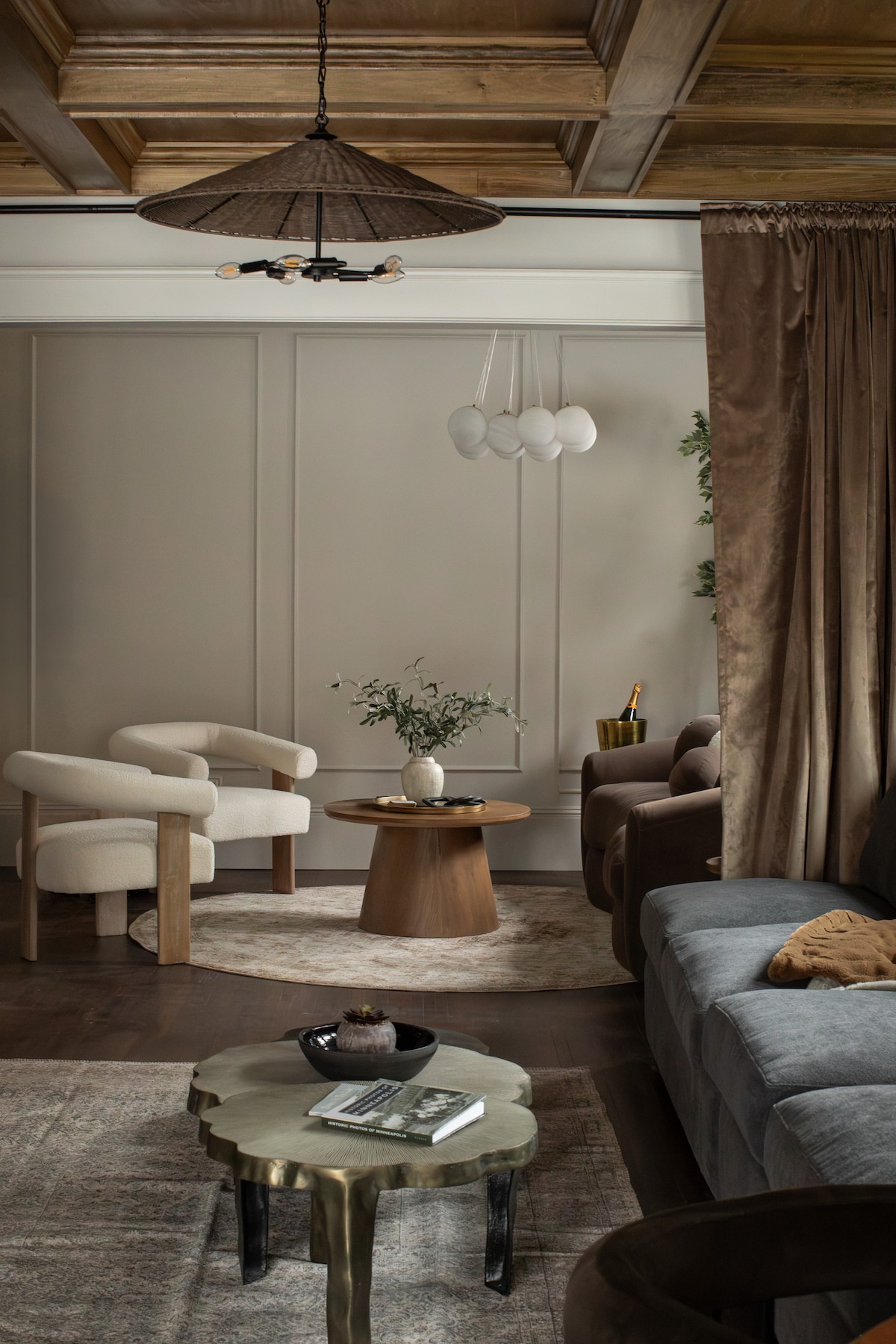 The living room showcases a blend of soft, neutral tones and textured materials. A plush sofa is complemented by two upholstered armchairs, with a circular coffee table at the center. A subtle area rug anchors the space beneath a coffered wood ceiling, illuminated by a pendant light.