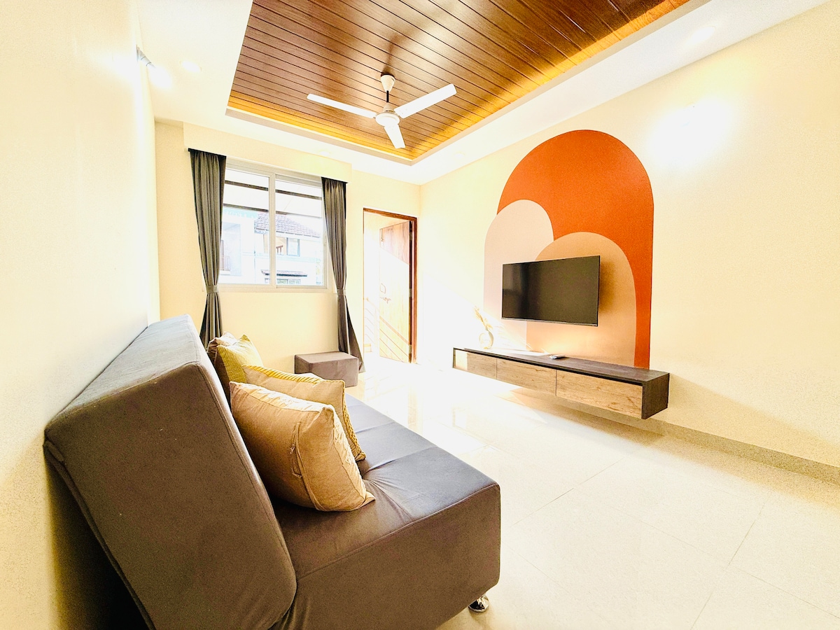 A spacious living area is presented with a modern couch and accent pillows. A flat-screen TV is mounted on the wall above a sleek wooden console. Natural light enters through a large window, while a ceiling fan provides airflow. The walls feature a tasteful design with earthy tones.