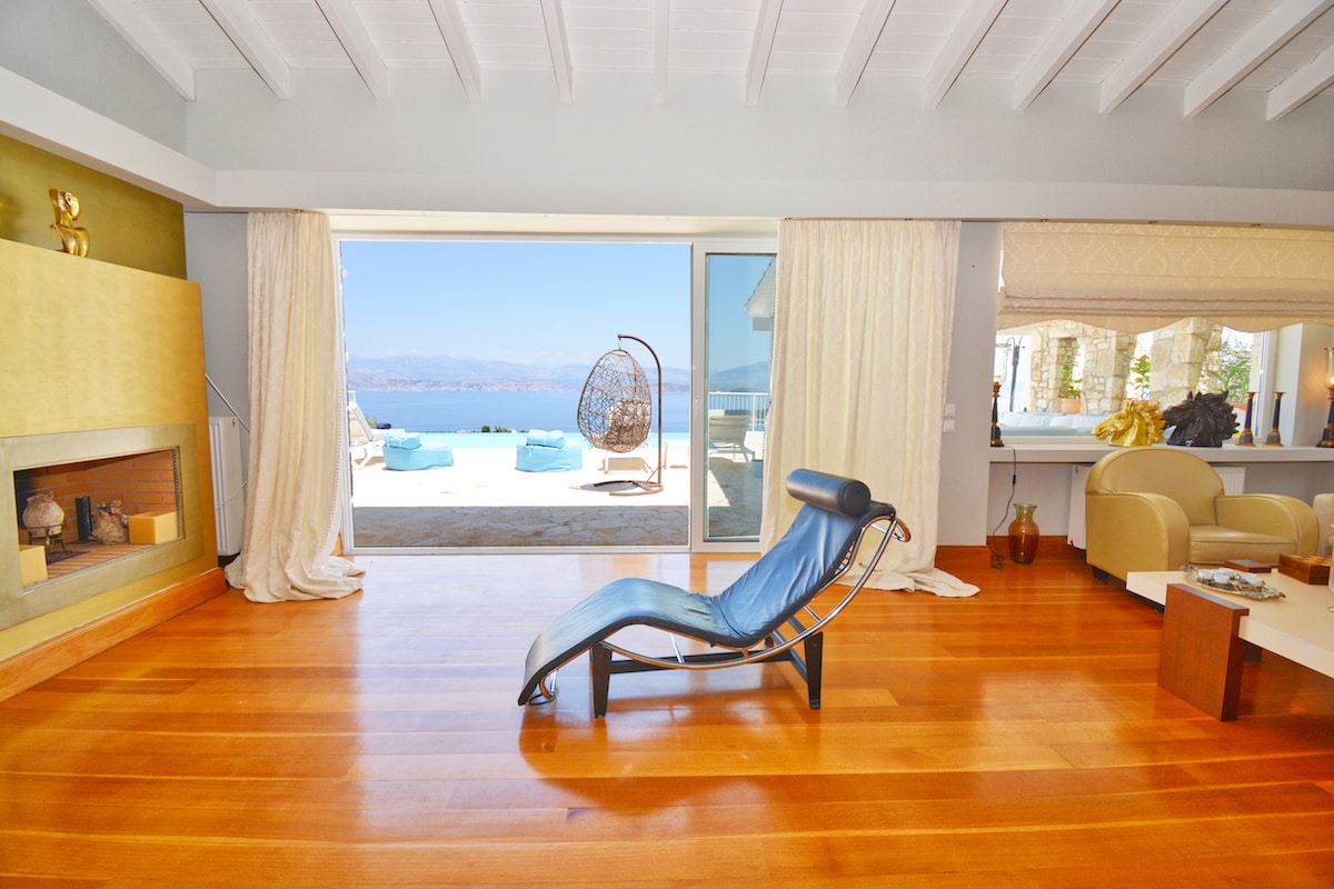 A spacious sitting room features a leather lounge chair positioned near large glass doors that open to a terrace with an unobstructed sea view. Light wooden flooring complements the neutral-colored walls, while soft curtains frame the openings, allowing natural light to illuminate the space.