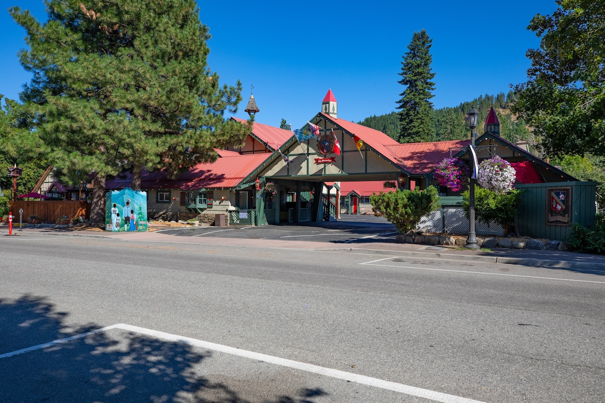 The exterior of the boutique motel is showcased, featuring a charming building with a red roof and whimsical architectural elements. Surrounding greenery includes tall trees and vibrant flowers. A winding road and parking area are visible in front of the entrance, enhancing accessibility for visitors.