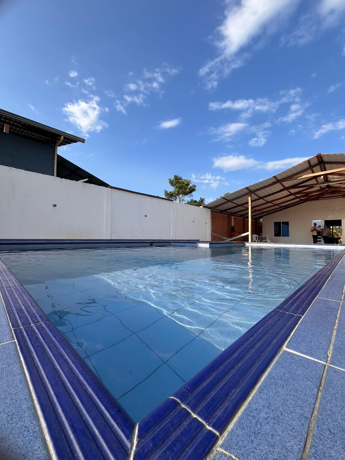 A clear blue pool is surrounded by a white wall and a shaded area with a ceiling overhead. The tranquil water reflects the bright sky, which is dotted with a few clouds. Lounge chairs are positioned near the pool, inviting relaxation.