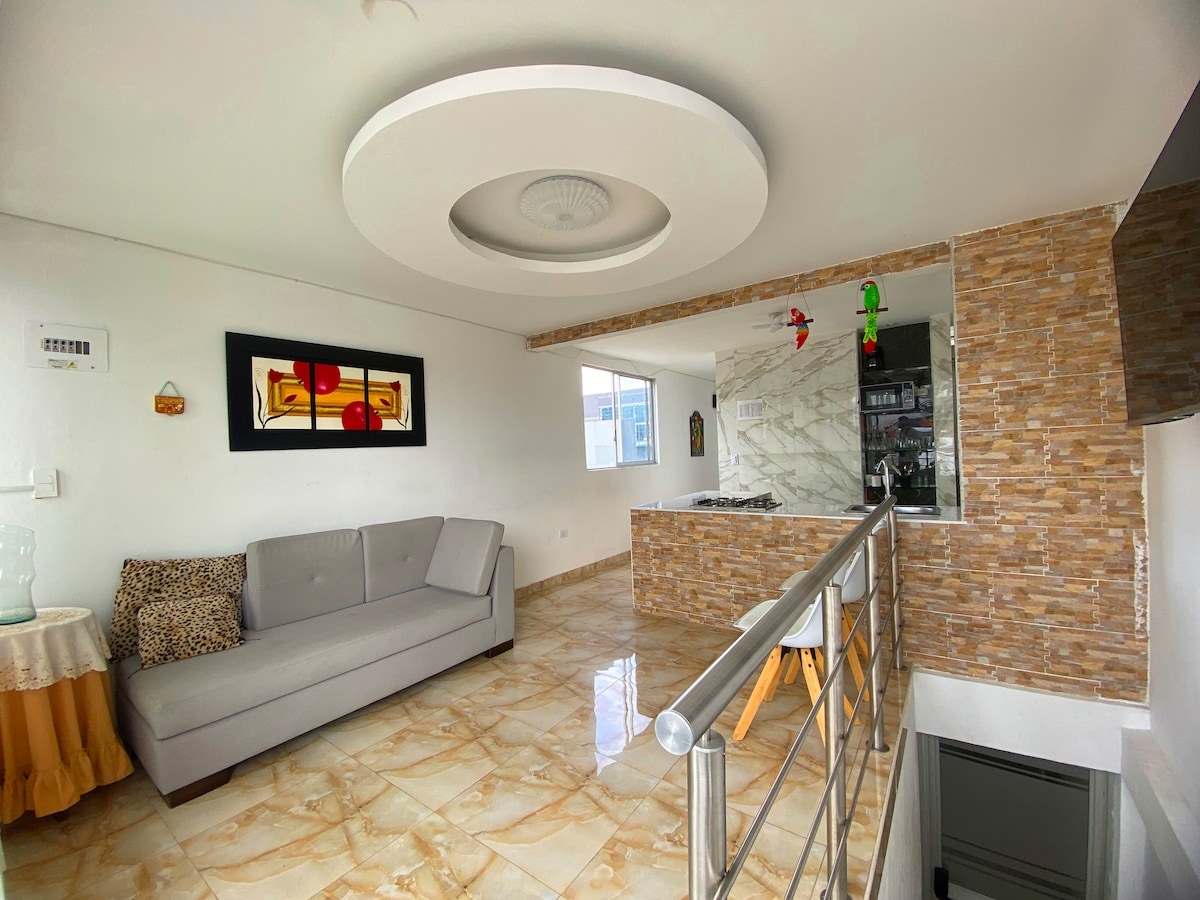 A modern living area is presented, featuring a light gray sofa against a white wall. Artwork is displayed above a stylish bar, with a marble countertop visible in the background. Natural light illuminates the space, accentuating the light-colored tiled flooring.