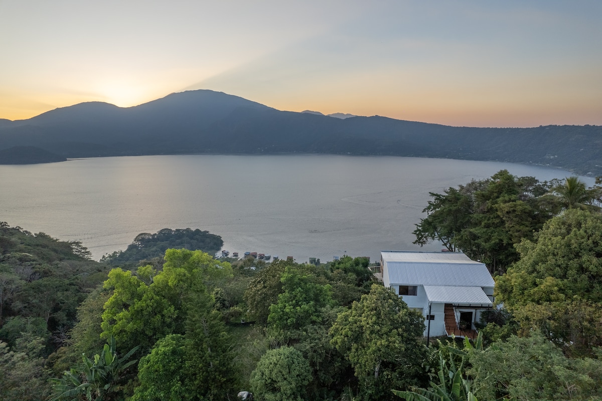The image captures a panoramic view of Lake Coatepeque at sunset, framed by lush greenery and distant mountains. The two-level house is visible in the foreground, surrounded by tropical foliage, emphasizing its serene hilltop location.