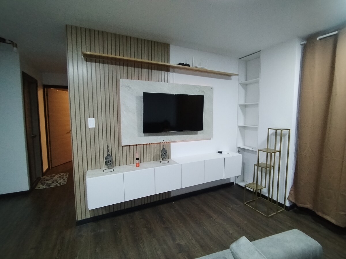 A modern living area is presented with a flat-screen television mounted on a textured wall. Below, a sleek white media console provides storage, while wooden accents add warmth to the space. Tall, gold shelving units stand in the corner, and light-colored curtains frame the window.