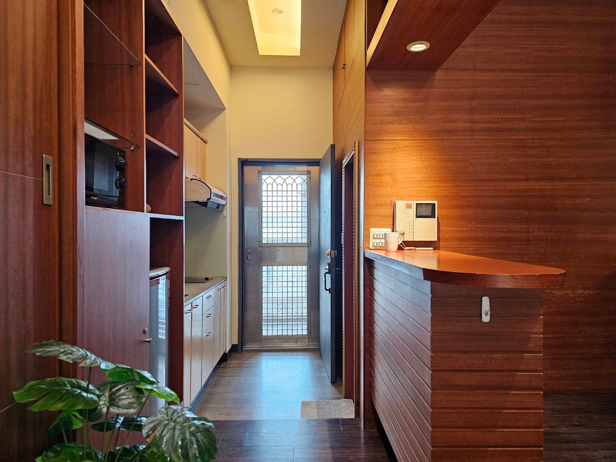 The entrance area displays wooden paneling with built-in shelves, housing a microwave and refrigerator. A door with a decorative grille leads outside, while a reception counter is positioned to one side. A potted plant adds a touch of greenery to the space.