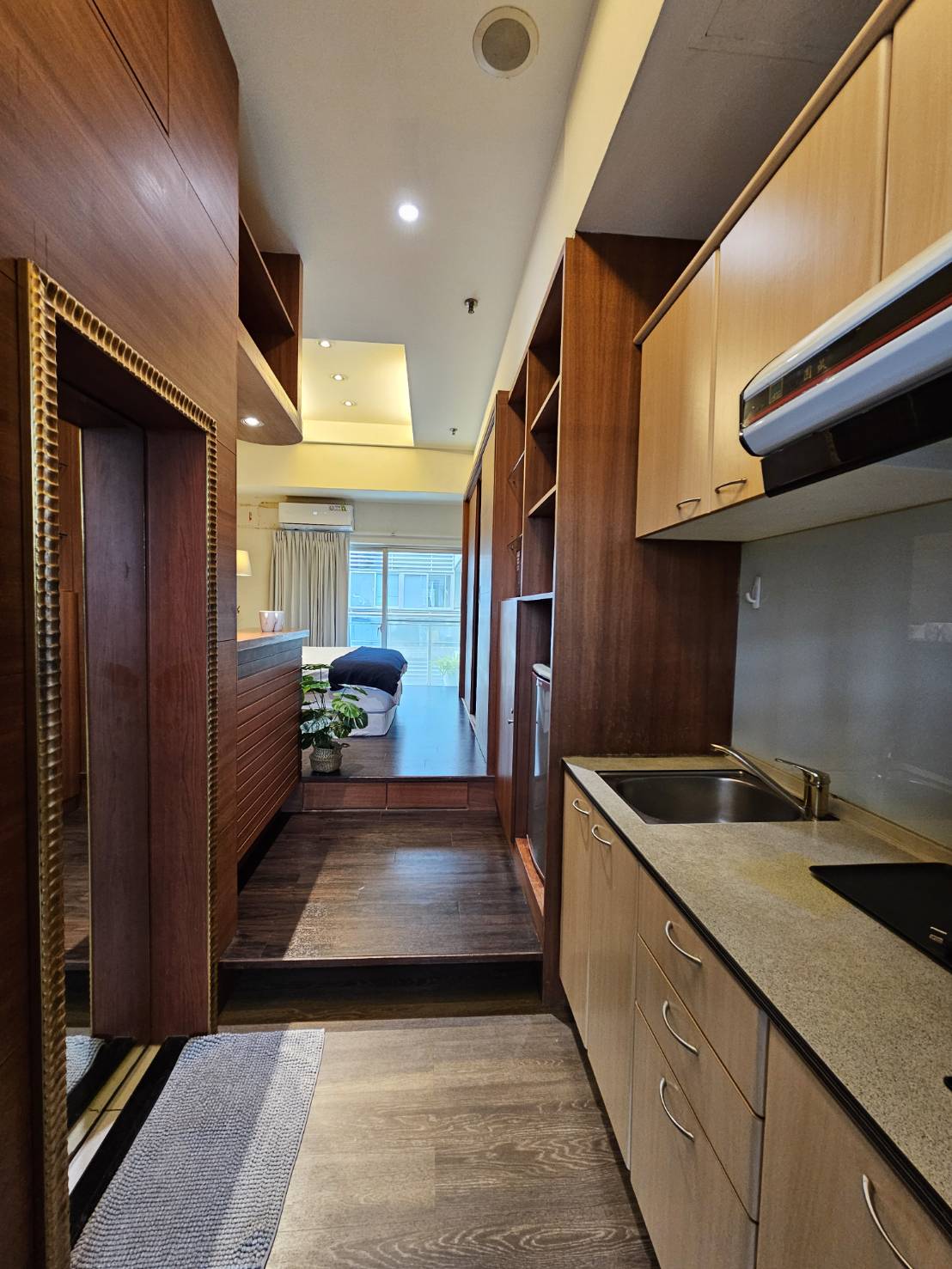 A modern kitchen area is featured with wooden cabinetry and granite countertops. It includes an integrated stovetop and a sink. Open shelving adds additional storage, while a plush rug lies at the entrance. Soft lighting creates a welcoming ambiance in the space leading to the bedroom.
