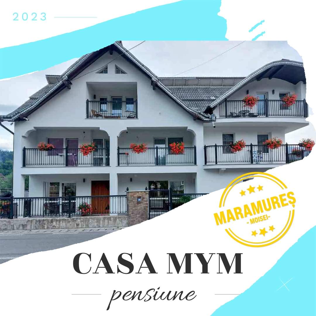 The exterior of Casa MYM exhibits a charming three-story building with a sloped roof. Balconies adorned with red flowers provide a warm touch. The main entrance is framed by a stone wall, and the design reflects a welcoming atmosphere in a natural setting.