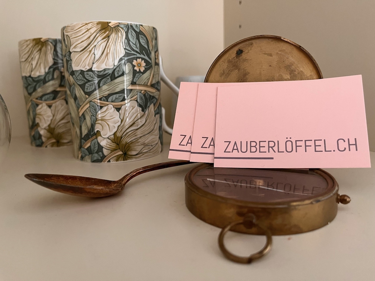 A decorative arrangement features two floral-patterned cups set in the background. In the foreground, a vintage brass spoon rests beside a compact mirror, which is opened to reveal its reflective surface. Several pink business cards are neatly placed beside the mirror.