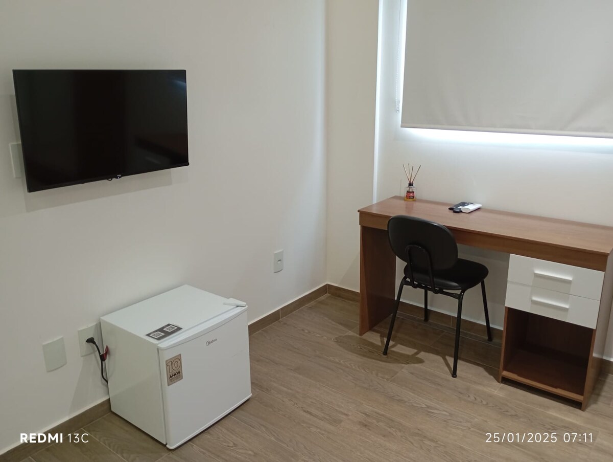 A compact room features a mounted flat-screen TV on the wall opposite a wooden desk with a chair. A small refrigerator sits on the floor, while a window allows natural light to illuminate the space. Neutral-colored walls contribute to a clean and organized look.
