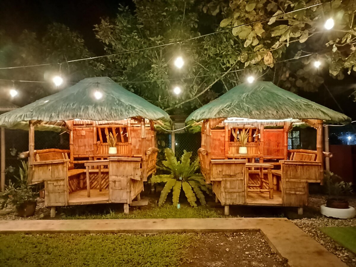 Two rustic kubos with thatched roofs are set in a lush outdoor area, surrounded by greenery. Soft lighting from strung bulbs highlights the inviting space, which features wooden seating arrangements within each hut, offering a cozy atmosphere for gatherings.