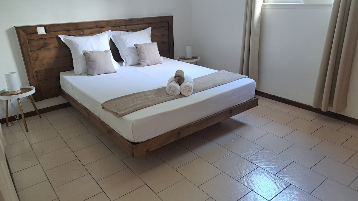 A spacious bed is centered against a wooden headboard in a well-lit room. Neutral-colored pillows are arranged on the bed. Two small tables with lamps are positioned on either side, and light curtains frame the window, allowing natural light to enhance the serene atmosphere.