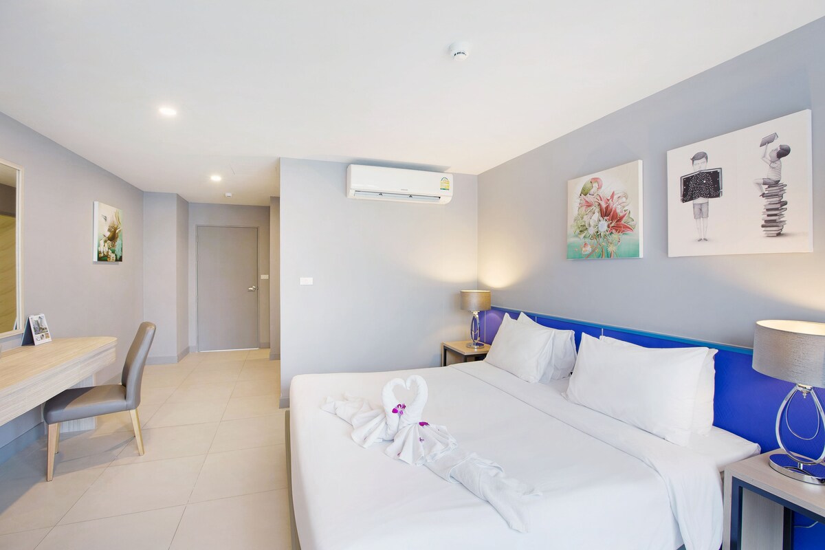 A modern guest room features a spacious bed adorned with white linens. Decorative wall art adds a touch of style, and a desk with a chair is positioned nearby. Soft lighting from the bedside lamps creates a calm ambiance in the sleek, minimalist space.