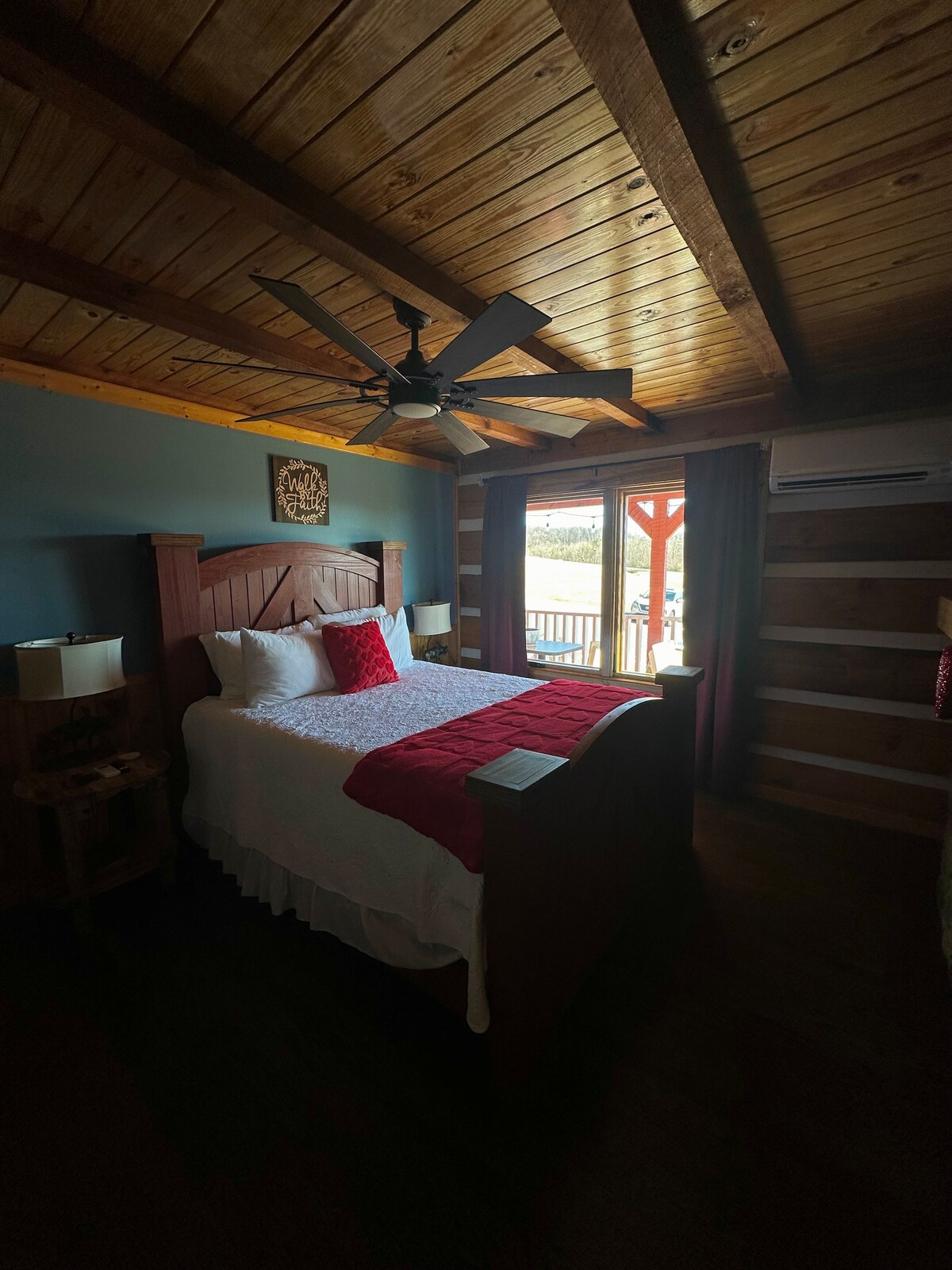 A cozy bedroom features wooden beams across the ceiling and a spacious bed dressed in white linens with a deep red accent blanket. Natural light enters through a large window, illuminating the room. Nearby bedside lamps provide additional soft lighting.