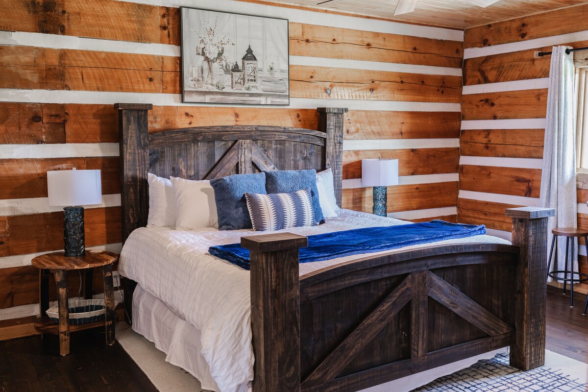 A rustic bedroom features a large wooden bed with a textured blue blanket and decorative pillows. Flanking the bed are two bedside tables with lamps. The room’s wooden walls add warmth, complemented by a framed nautical artwork hanging above the bed.