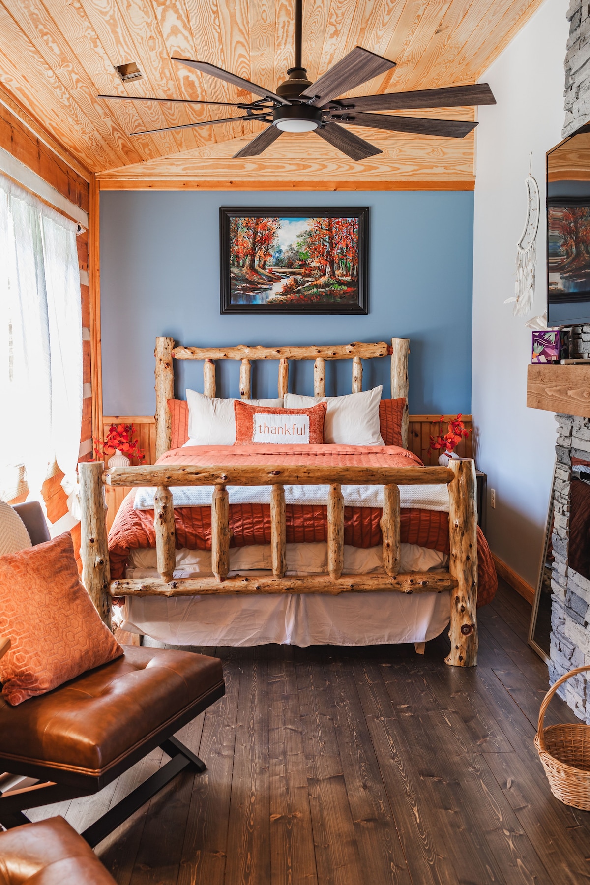 A rustic bedroom features a unique log bed with a terracotta bedding set. A ceiling fan hangs above, and an artwork depicting a forest scene is mounted on the blue wall. An armchair sits nearby, complemented by warm decorative cushions.