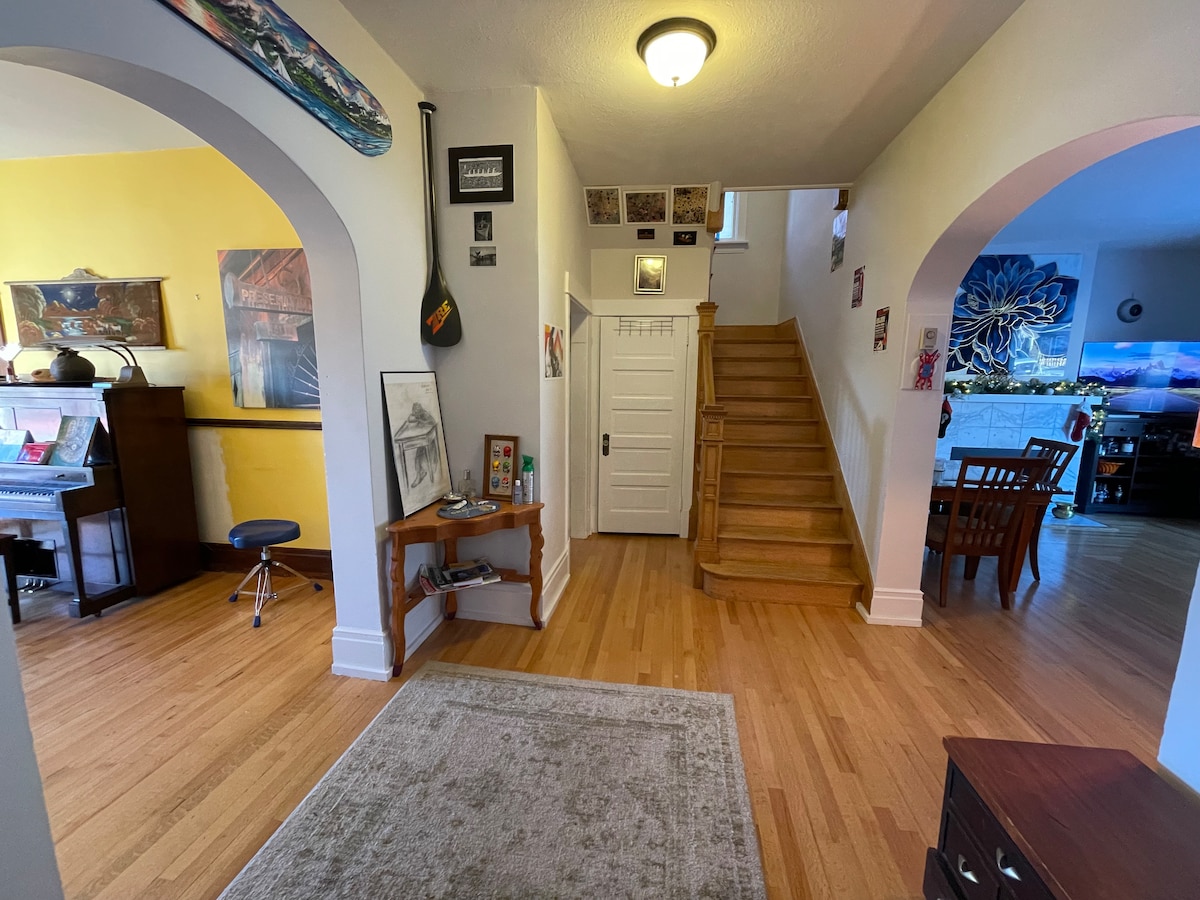 An inviting entryway is highlighted by archways leading to a spacious living area and staircase. A wooden side table displays framed artwork, while a piano is positioned nearby. Light-colored walls provide a bright contrast to the warm wooden flooring.