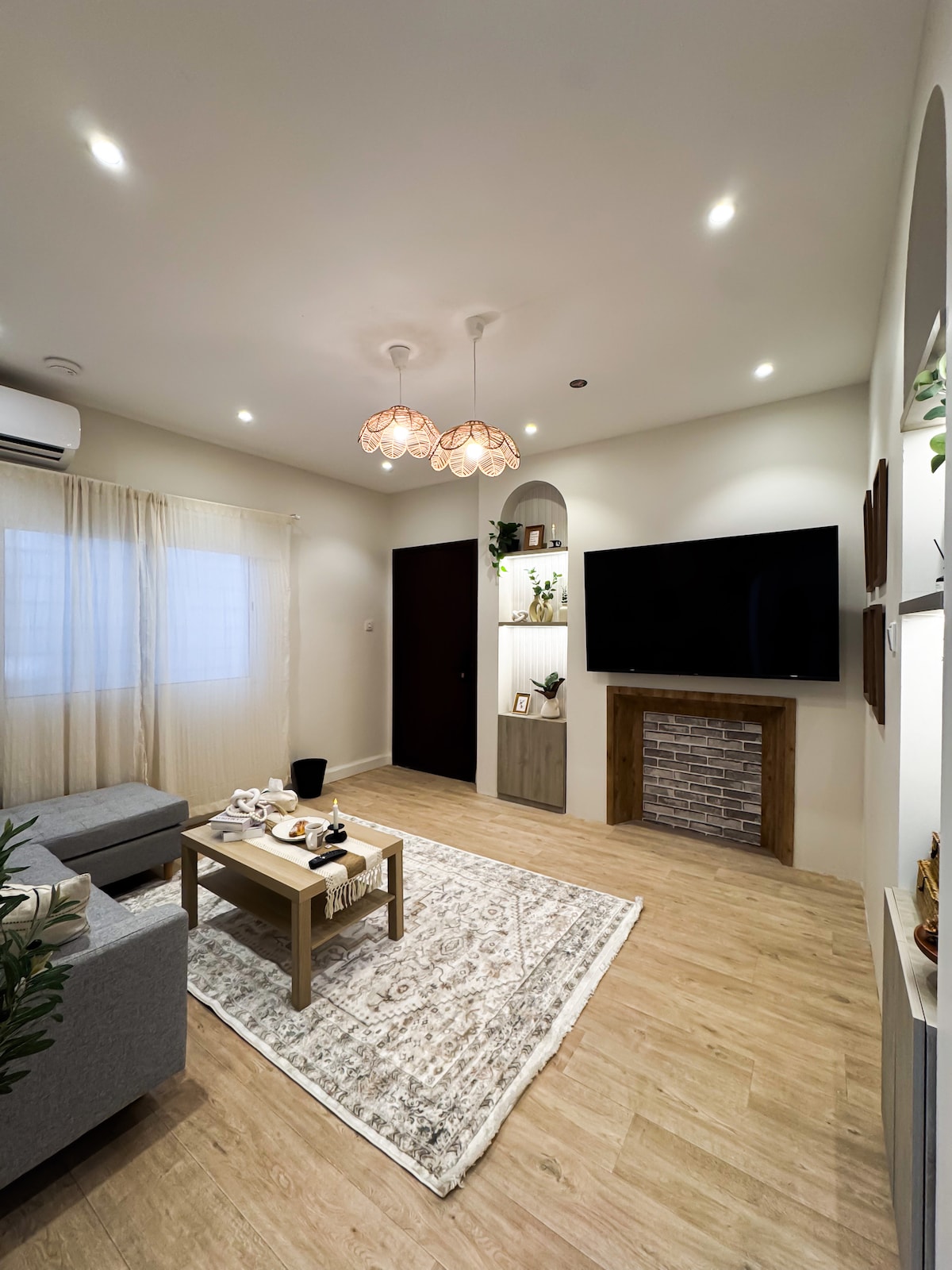 A stylish living room is showcased, featuring a large flat-screen TV mounted on the wall. A light-colored rug covers the wooden floor, and a small coffee table holds decorative items. Soft lighting is provided by two pendant lamps, and a cozy seating area is included.