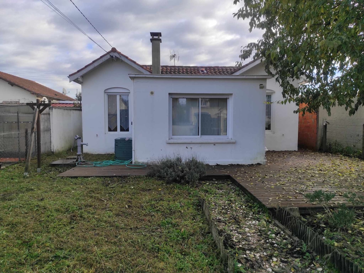 A charming white house is presented with a wooden terrace and a spacious grassy yard filled with scattered leaves. Large windows provide natural light, and a garden hose is seen next to a water tap. A serene outdoor environment is visible in a peaceful setting.
