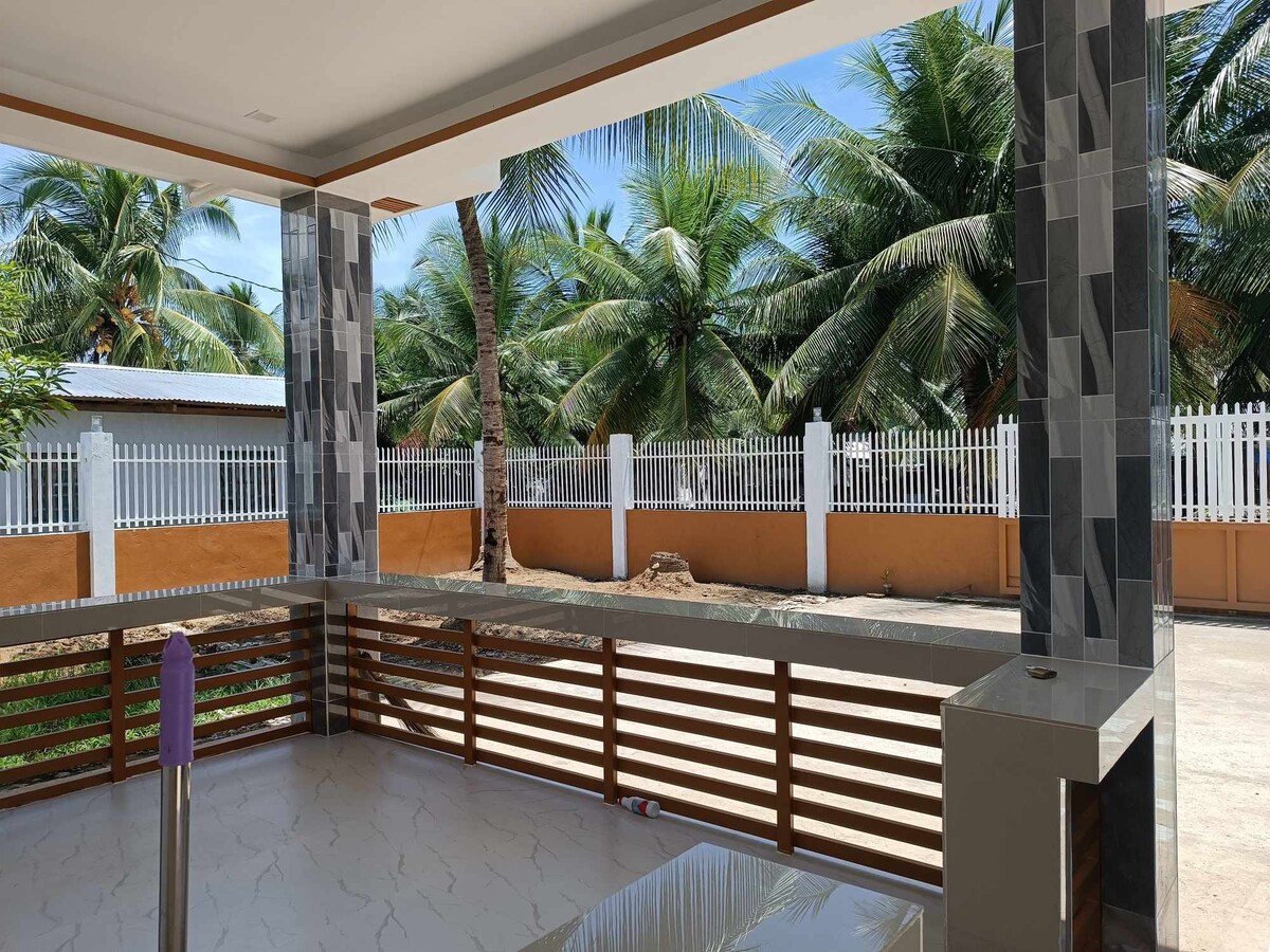 A spacious outdoor patio is surrounded by tall palm trees, creating a serene environment. The area features a tiled floor and modern wooden railings. Natural light illuminates the space, and a glimpse of a fenced property is visible in the background.