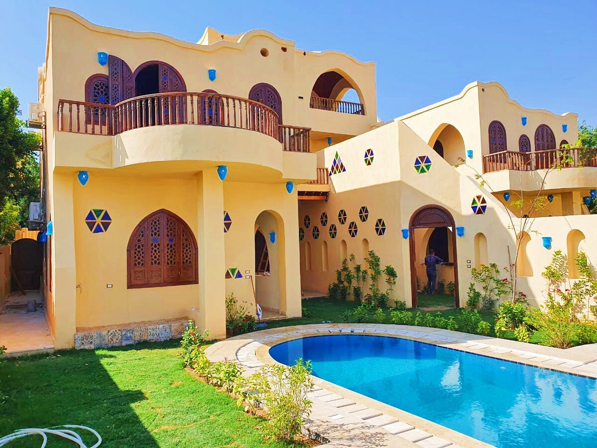 The villa's exterior showcases vibrant colors and unique architectural details. A private swimming pool is framed by a stone pathway, surrounded by a well-maintained garden. Balconies are visible on the upper level, featuring decorative railings that enhance the overall aesthetic.