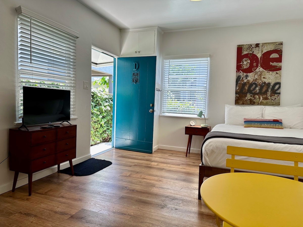 A bright studio apartment features a vibrant blue front door and large windows, allowing natural light to fill the space. A comfortable bed is positioned beside a stylish dresser. A small yellow table adds a pop of color, complementing the neutral wood flooring.