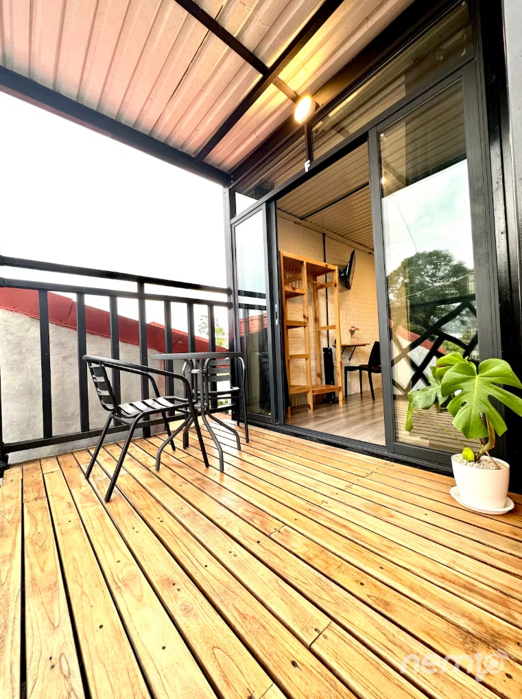 A private balcony features wooden decking with a small round table and two black chairs positioned beneath an overhang. A potted plant adds a touch of greenery. Large glass doors offer a view into the adjoining room filled with natural light.
